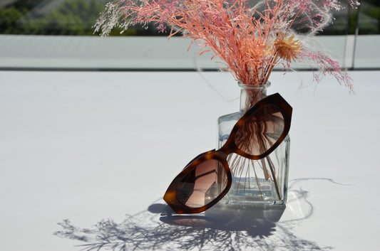 A pair of Merhoki Havana Brown sunglasses resting against a small glass vase filled with pink and yellow dried flowers. The sunglasses cast a soft shadow on the white surface, complementing the delicate floral arrangement.