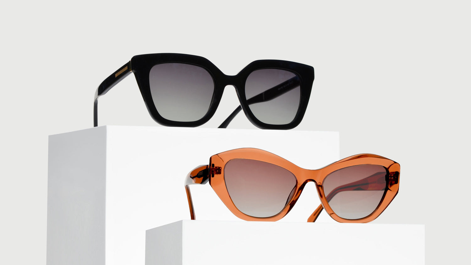 A pair of stylish cat eye glasses on white pedestals, the top features a black square frame, the bottom has a translucent pink frame for a youthful look.