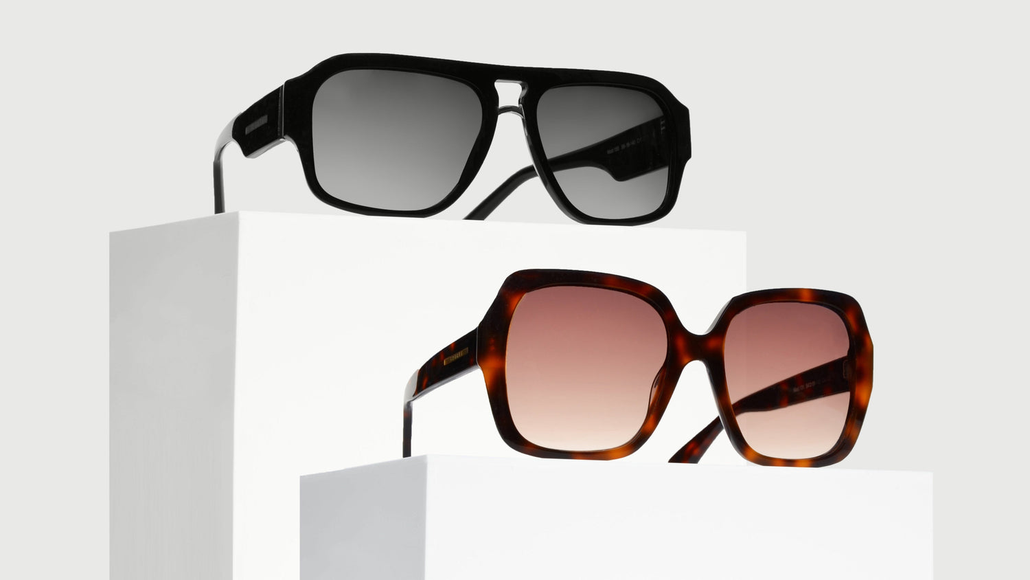A pair of oversized bold eyewear on white pedestals. The top pair is a black double bridge navigator, the bottom is a Havana brown square butterfly frame.