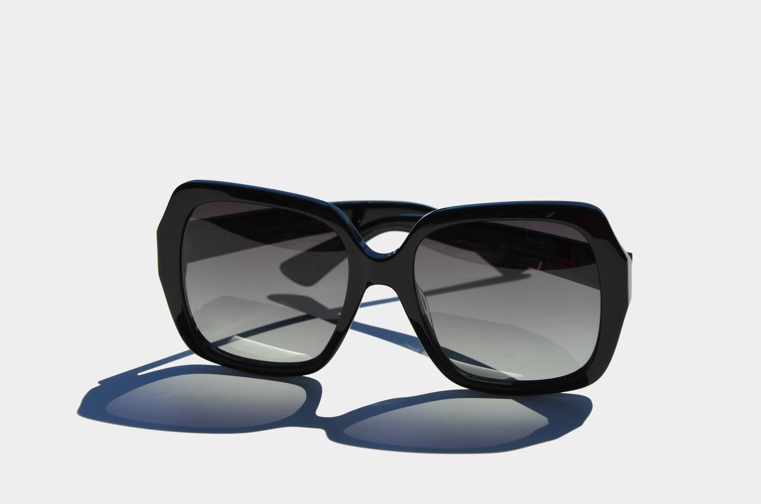 Black oversized sunglasses with grey lenses, shown upright on a flat surface with subtle shadows and a bold, angular design