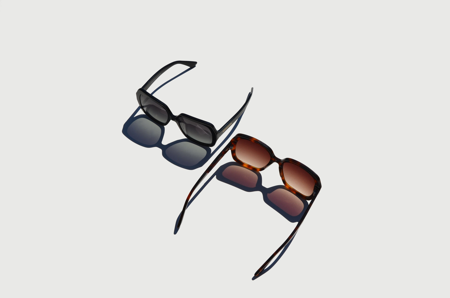 Two pairs of sunglasses, one black with gray lenses and the other havana brown with amber lenses, shown from above with their arms extended and casting shadows.