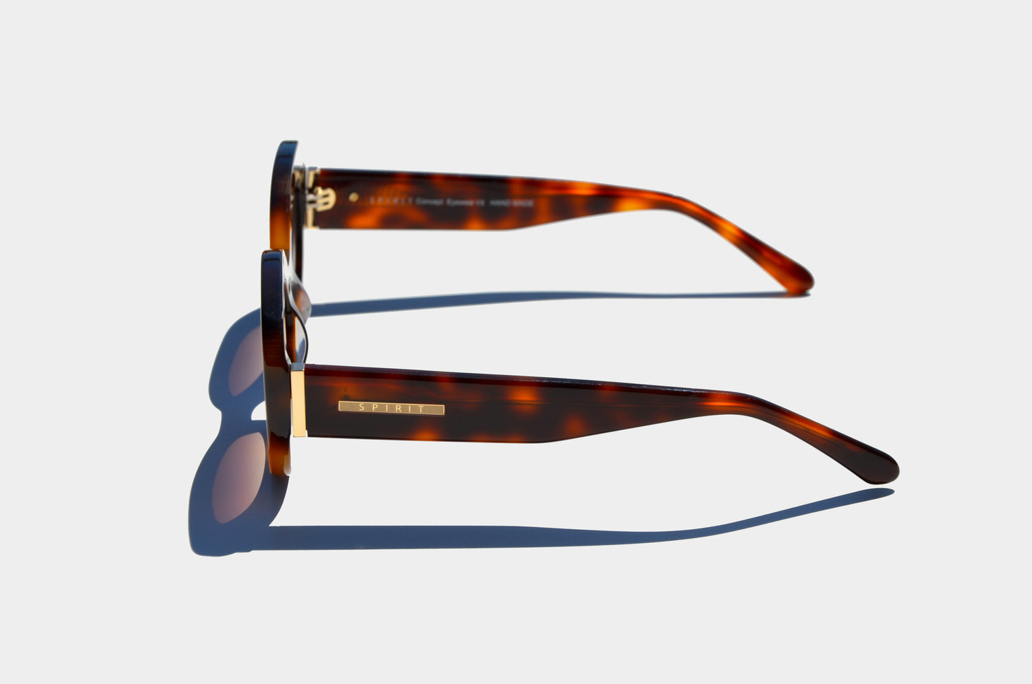 Brown sunglasses from the side, showcasing extended arms with a gold “SPIRIT” logo engraved. The gradient brown lenses cast shadows beneath.