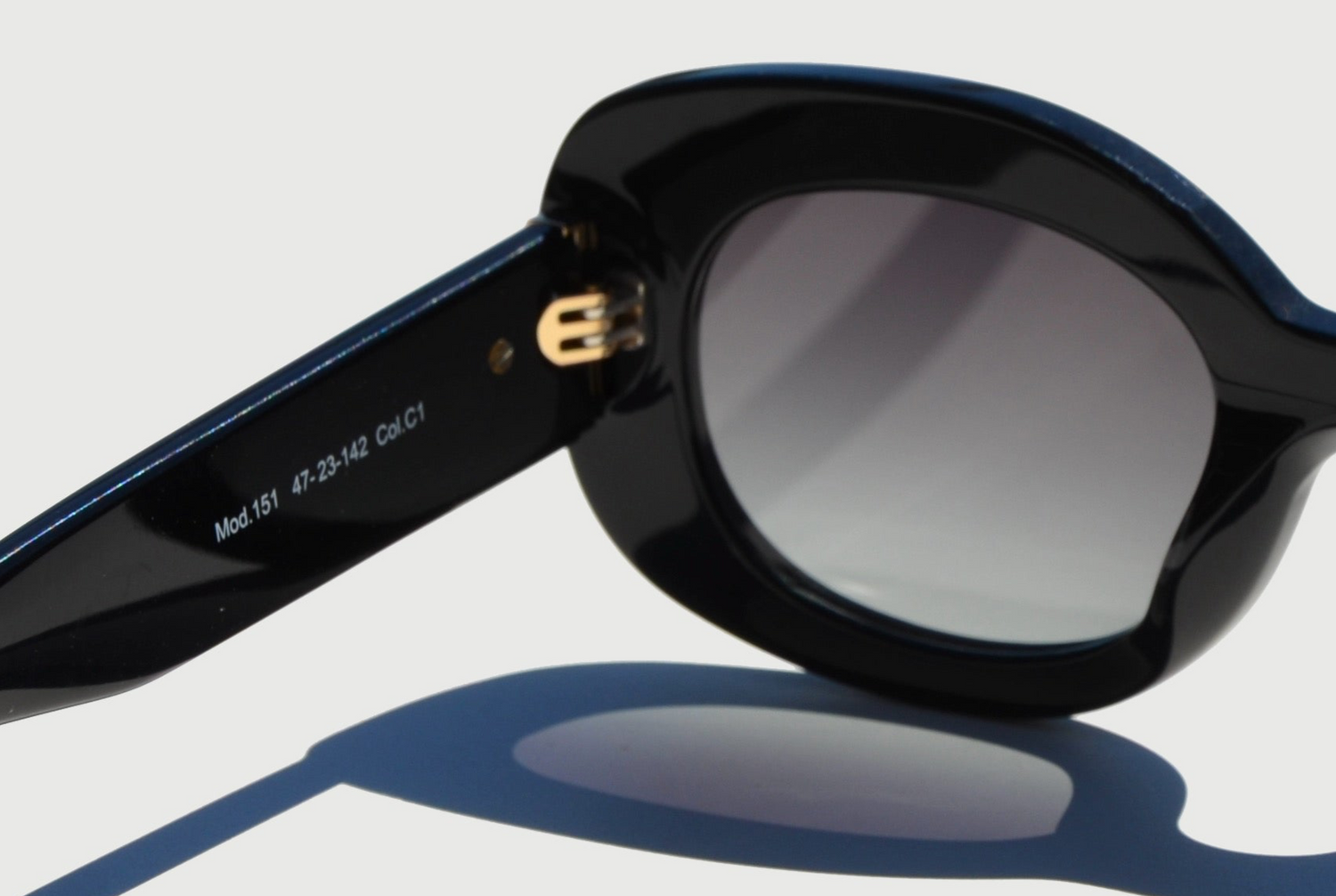 Close up thick black sunglasses oval grey lens, gold hinge, and arm extended out