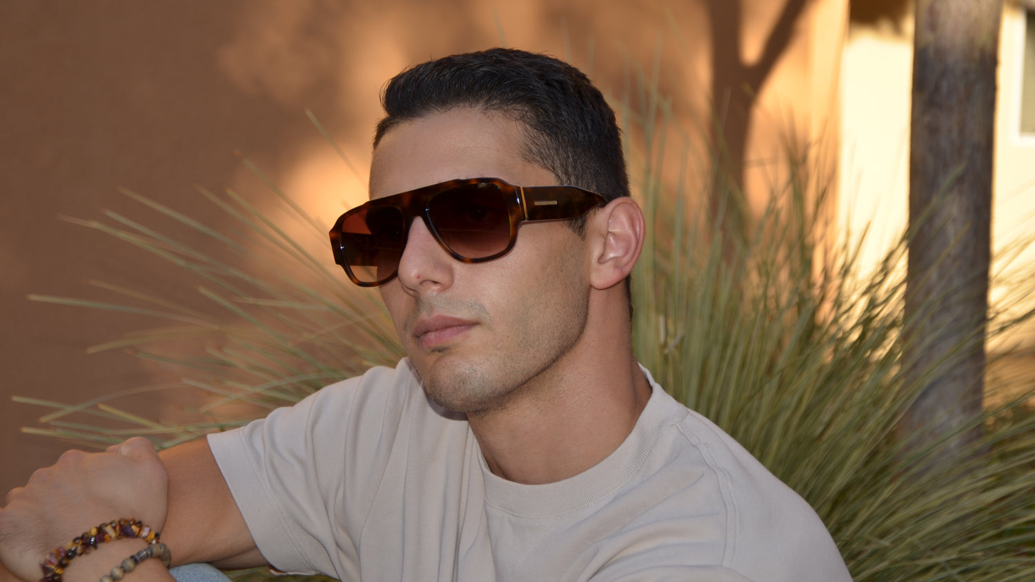 Man posing hands wrapped around bent leg wearing brown sunglasses head tilted to side