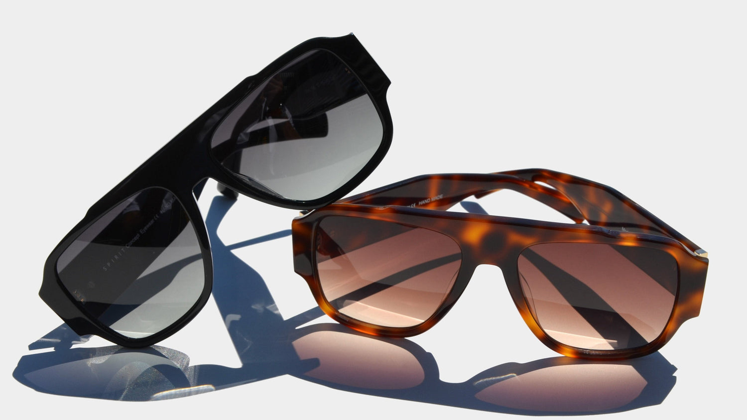 A sleek presentation featuring two shield style eyewear pieces on a minimalistic light background. On the left, a pair of black shield eyewear with gradient gray lenses is propped slightly upward. On the right, a Havana brown frame with warm amber-tinted lenses stands upright.