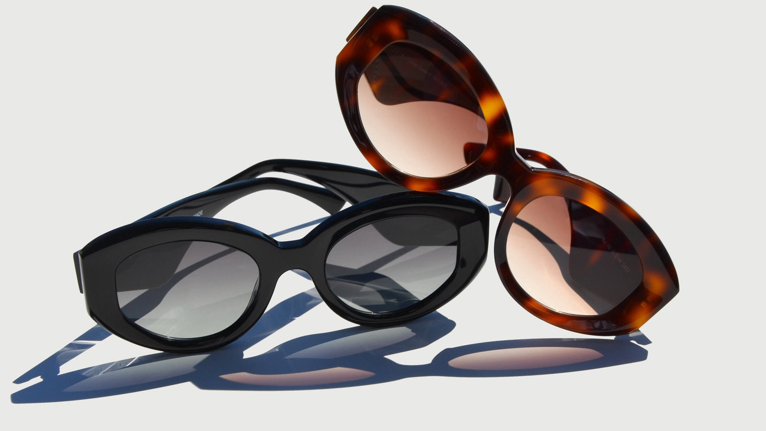 A stylish arrangement of two oval eyewear pairs displayed on a light background. The foreground showcases a black pair with dark gradient lenses, exuding modern elegance. Layered behind, the Havana pair features a brown frame with warm amber-tinted lenses, offering a bold and classic appeal. 