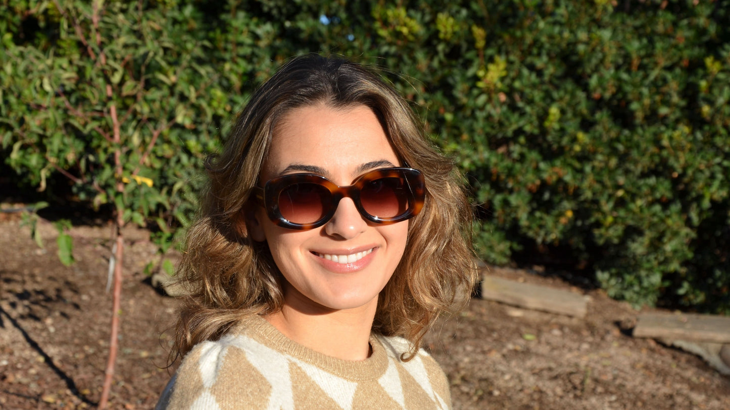 Woman wearing Jacky rounded cat-eye sunglasses in havana brown, smiling brightly, styled with a beige diamond-pattern sweater amidst an autumn landscape.