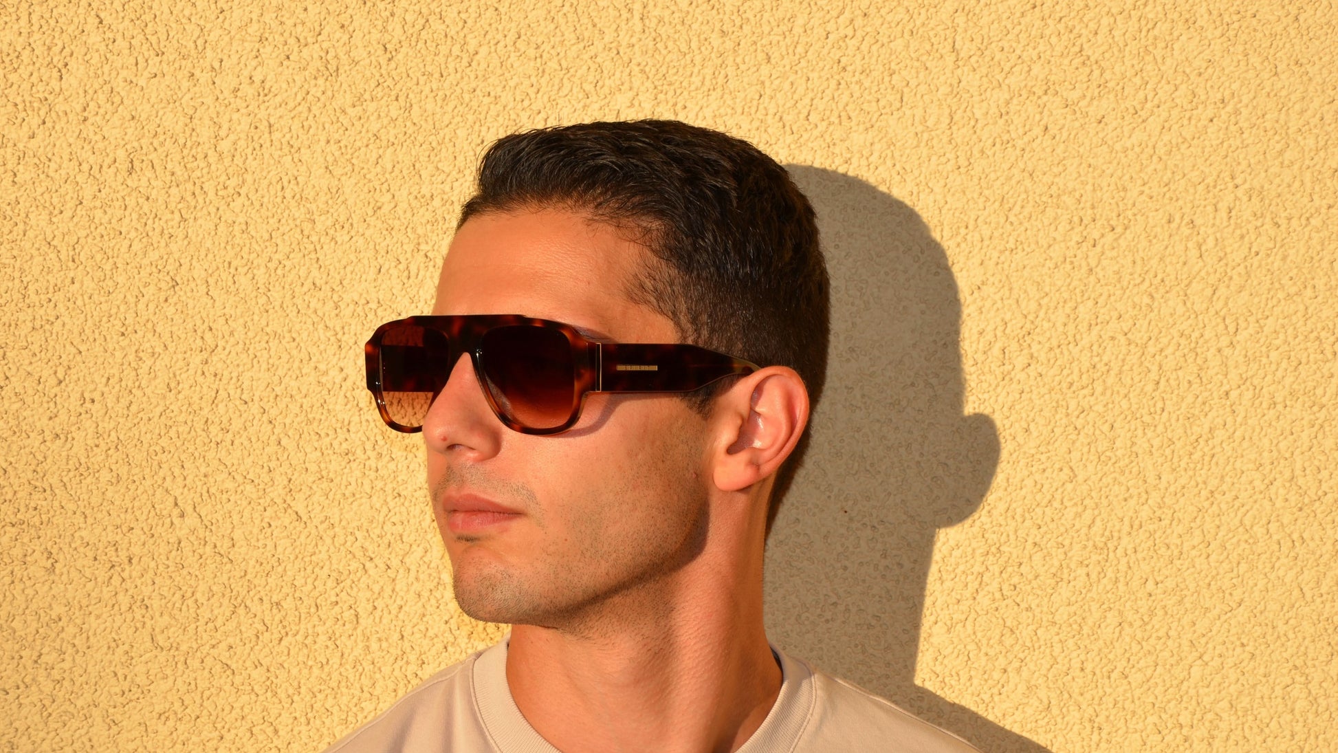 A man, head turned slightly to the side, wearing Havana rectangular navigator eyewear poses against a sunlit textured yellow wall. 