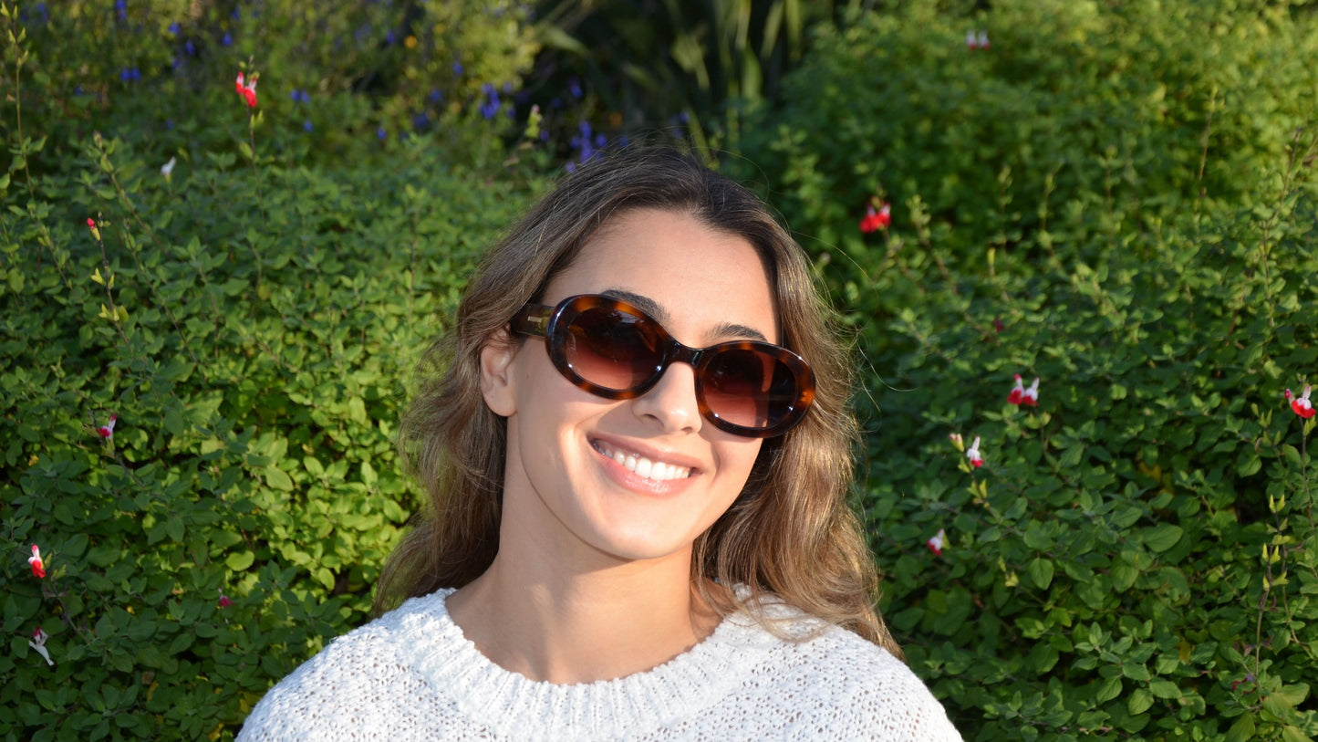 A smiling woman wearing Riley oval havana brown sunglasses with sleek frames and gradient lenses, posing outdoors against a vibrant green garden with blooming flowers, dressed in a cozy white knit sweater.