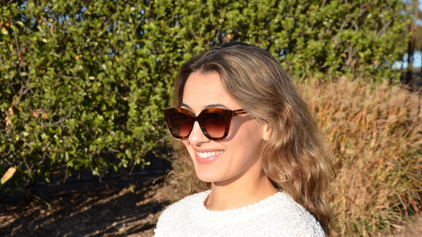 A smiling woman wearing Robin square cat eye Havana brown sunglasses, featuring amber lenses and chic frame, set against a vibrant green foliage background under natural sunlight.