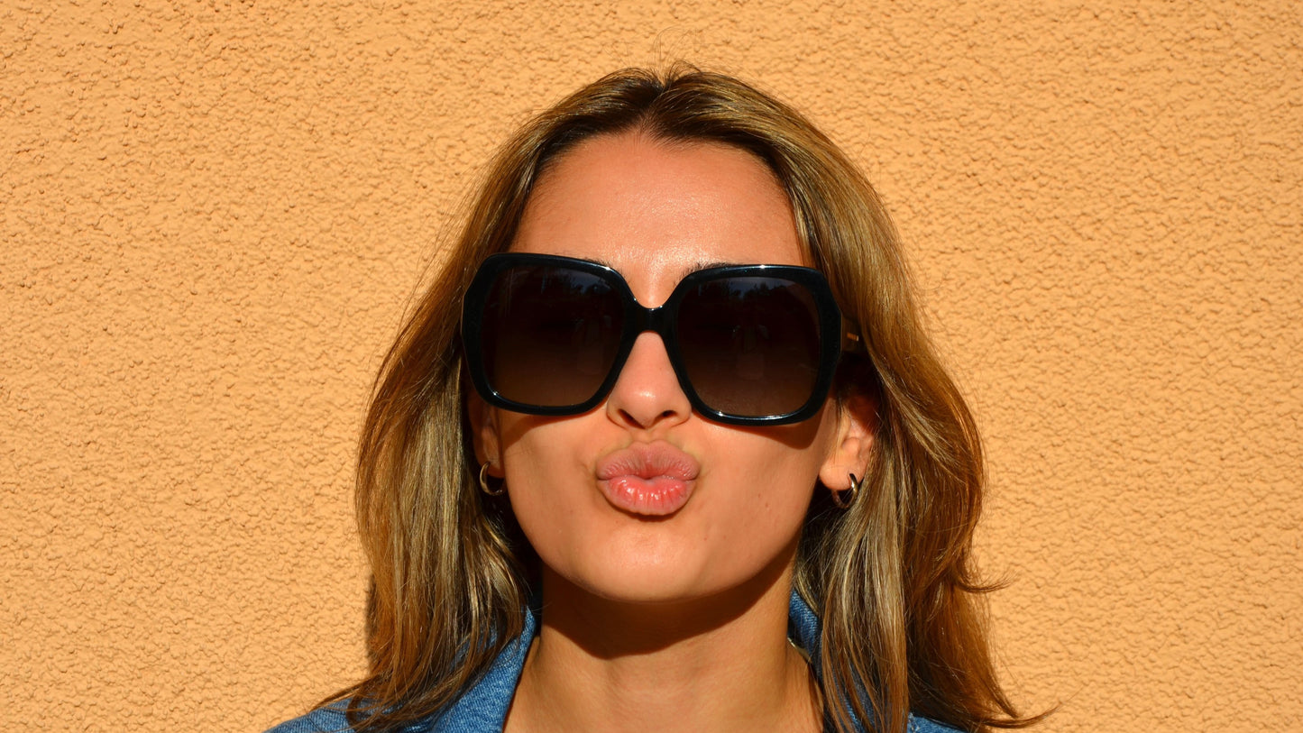 A woman wearing oversized black butterfly eyewear posing against a peach wall, making a playful kissing gesture, dressed in a denim jacket.