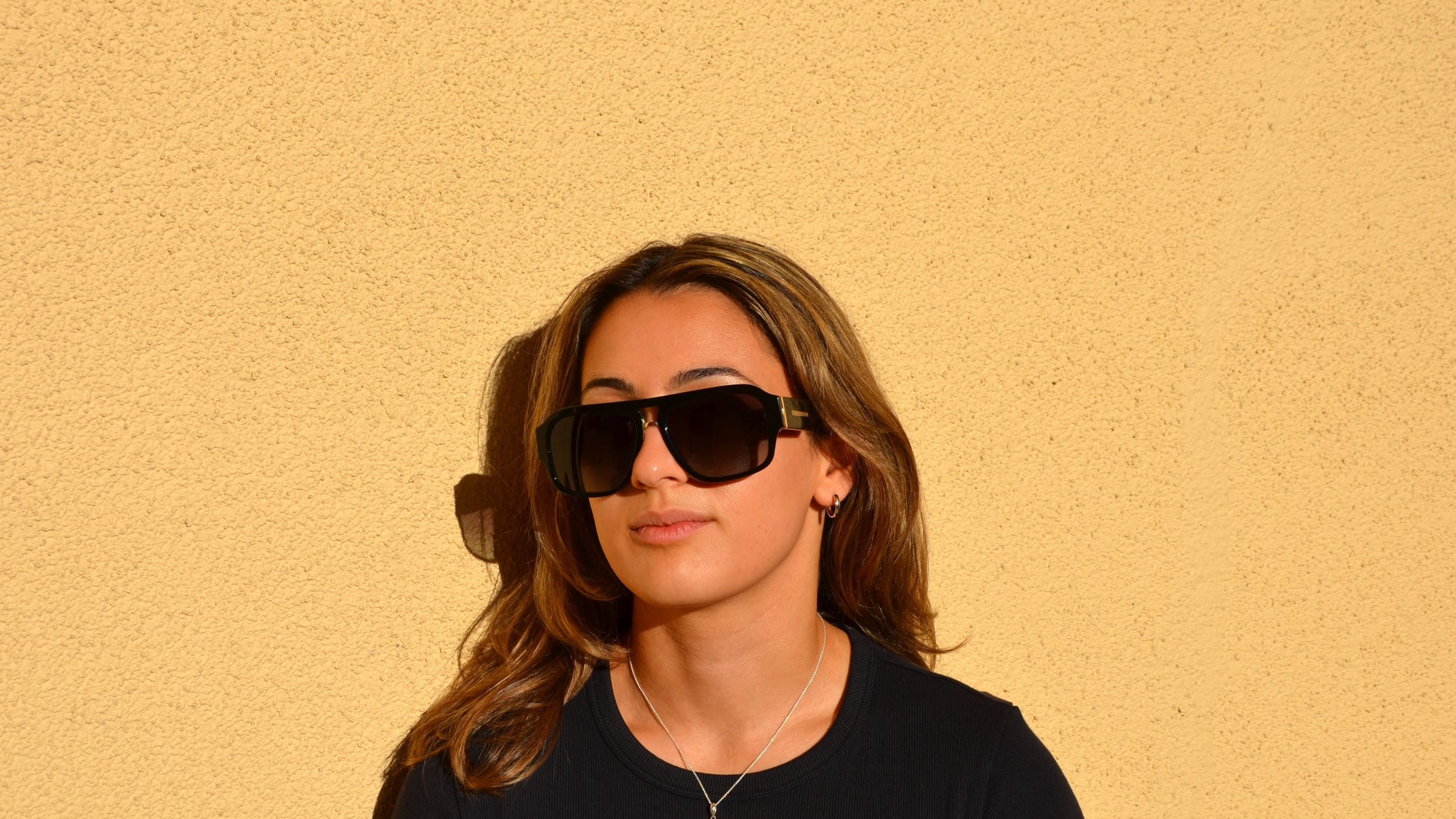 A woman wearing black navigator eyewear and a black shirt, standing against a yellow wall with subtle shadows and sunlight.