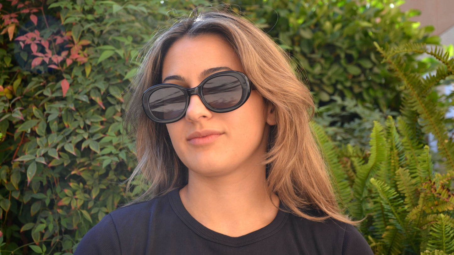 A woman wearing black oval eyewear, paired with a black shirt, posing in front of lush greenery under natural sunlight.