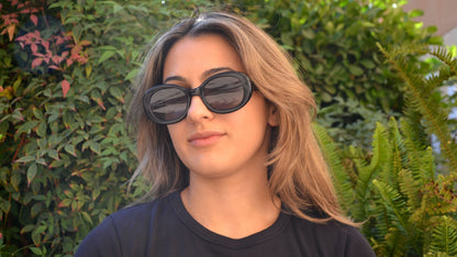 A woman wearing black oval eyewear, paired with a black shirt, posing in front of lush greenery under natural sunlight.