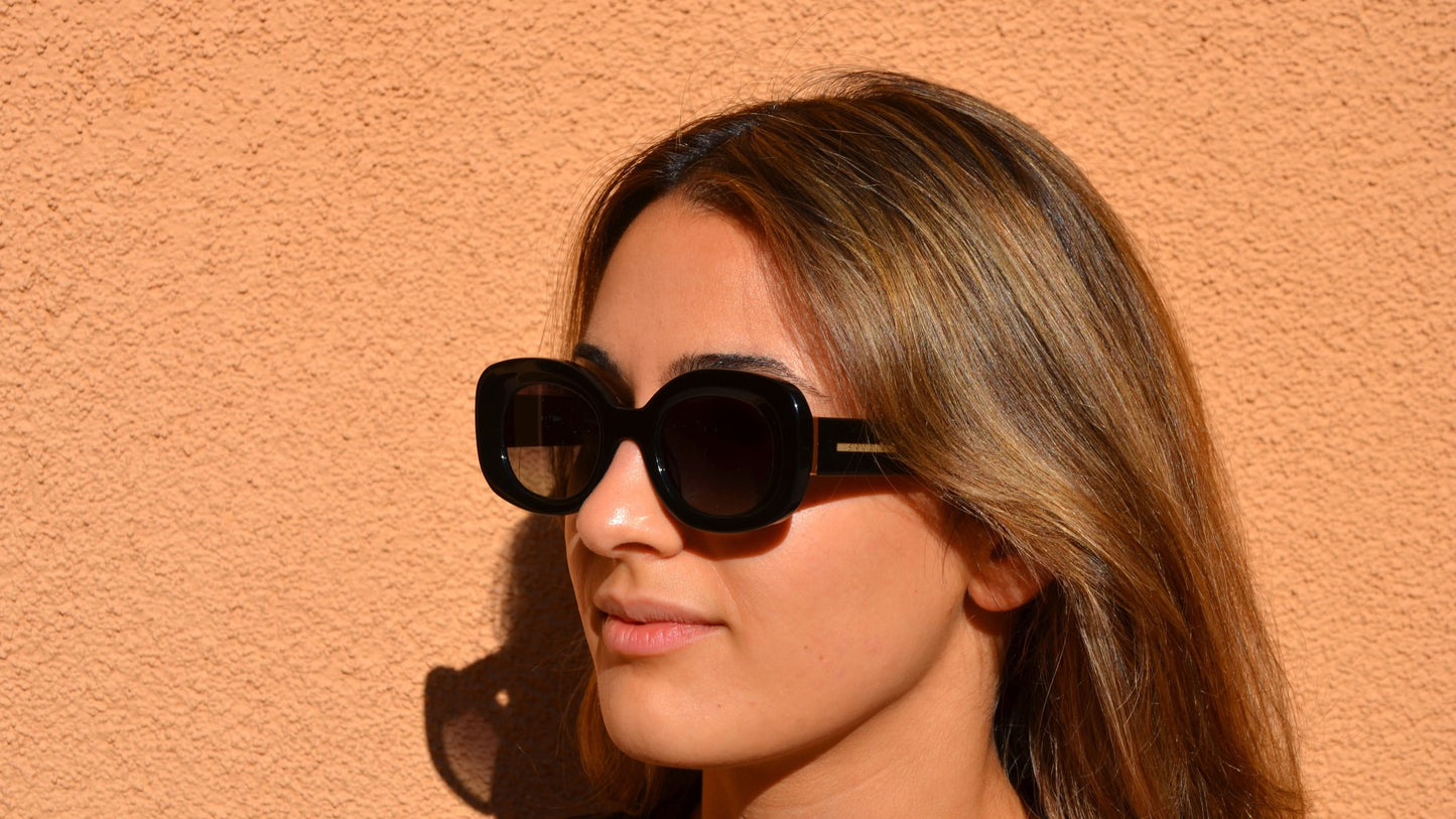 A woman wearing black rounded cat-eye eyewear, posing against a peach wall, with sunlight highlighting her hair and face.