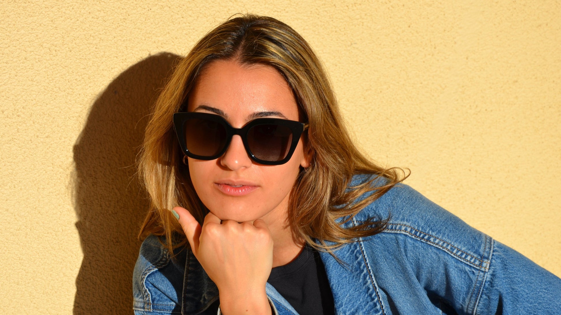 A woman wearing black square cat-eye eyewear, styled with a denim jacket and black top, posing against a yellow wall with sunlight casting shadows.