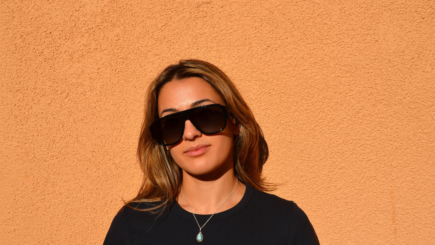 A woman wearing oversized black navigator eyewear with subtle golden accents stands in front of a textured orange wall. 